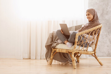 Young arabic woman in headscarf relaxing in wicker chair with laptop at home, watching movies or browsing internet, copy space