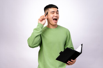 An Asian man in a green sweater is holding a black notebook and a pen while looking excited or...