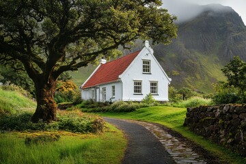 Fototapeta premium Charming white cottage with red roof beside winding path surrounded by greenery in a misty mountainous landscape during daylight