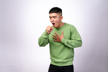 An Asian man in a green sweatshirt is coughing into his fist while placing his other hand on his back. His facial expression conveys discomfort. The background is plain white