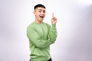 An excited Asian man in a green sweatshirt raises his index finger with an open-mouthed expression,...