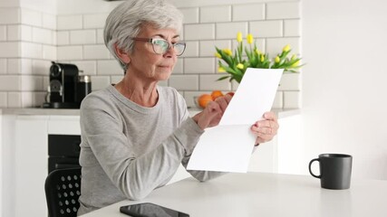 Elderly Lady Carefully Reading Financial Notice Letter In The Kitchen