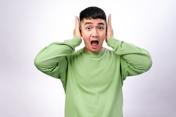 An Asian man in a green sweatshirt covers his ears with both hands while shouting. His eyes are...