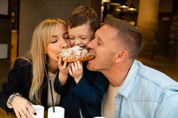 Mom dad and little son bite one French croissant. Happy family have tea party breakfast at a table...