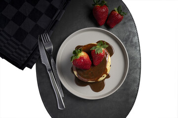 A stack of pancakes with chocolate sauce and strawberries on a gray plate, placed on a black leather mat with a fork and knife beside a checkered napkin.