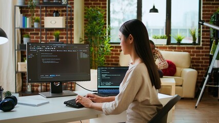 Asian programmer seen typing code for a user interface design on dual monitors, working in a...
