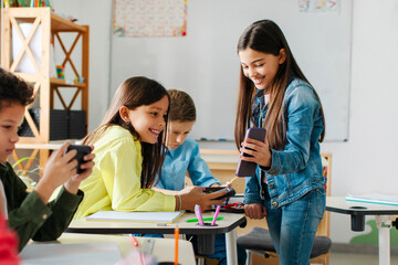 Break at school. Schoolgirl showing cellphone to friends, sitting in classroom, capturing the...