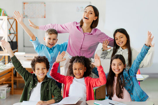 Excited teacher and group of diverse school classmates raising arms and smiling, sitting in classroom, celebrating the last day of school