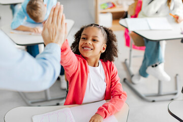 Little black schoolgirl giving high five to teacher and smiling. Students with goals, celebration at school desk