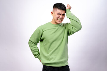 An Asian man in a green sweatshirt against a white background is grimacing while pressing his hand...