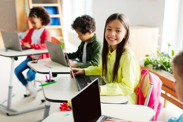 Happy schoolgirl sitting at lesson using laptop computer, learning online with classmates in modern classroom. Technology and education concept