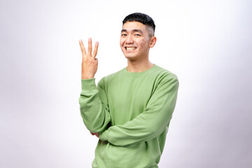 An Asian man in a green sweatshirt is smiling while holding up three fingers on his right hand. His left arm is crossed, and he stands against a white background, expressing confidence and enthusiasm