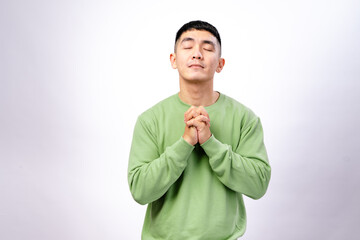 An Asian man in a green sweatshirt stands against a white background with his hands clasped together, and a calm expression, as if praying, hoping, or making a wish.