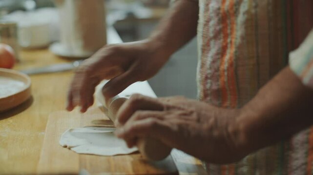Hands of man using rolling pin to flatten dough into thin round sheet on wooden table, preparing homemade Indian flatbread in kitchen. Close-up view