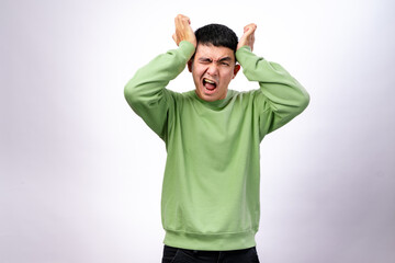 An Asian man wearing a green sweatshirt against a white background is clutching his head with both...