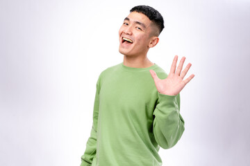 A cheerful Asian man in a green sweatshirt waves his hand with a bright smile. He stands against a...