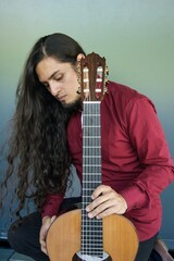 A long-haired guitarist in a red shirt leans on his classical guitar with a contemplative expression against a soft gradient background.