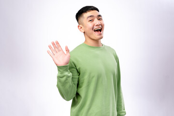 A cheerful Asian man in a green sweatshirt waves his hand with a bright smile. He stands against a...
