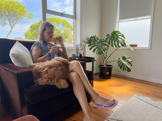 A lady in shorts sits in a chair eating with a green cheese plant in a room .Sat Beside her is a ginger coloured hairy dog.Outside the window is blue sky and trees.Homely scene.