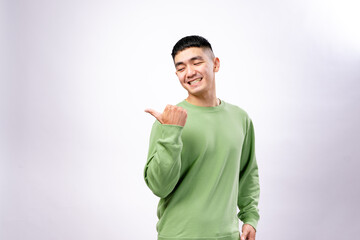 A cheerful Asian man in a green sweatshirt smiles playfully while pointing his thumb over his shoulder. He poses casually against a plain white background, creating a fun and friendly expression