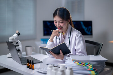 A woman in a white lab coat is sitting at a desk with a laptop and a notebook