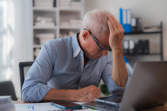 Senior businessman sitting at his office desk holding his head with his hand, showing concern and suffering from headache or migraine, having problems at work or from overworking