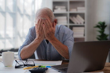 Senior businessman sitting at desk with laptop and paperwork is covering his face with his hands,...