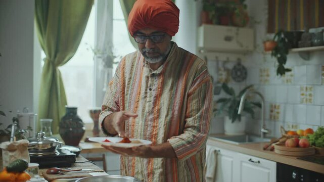 Indian chef in turban describing various spices on plate and explaining how to use them in cooking as giving online cooking class in kitchen