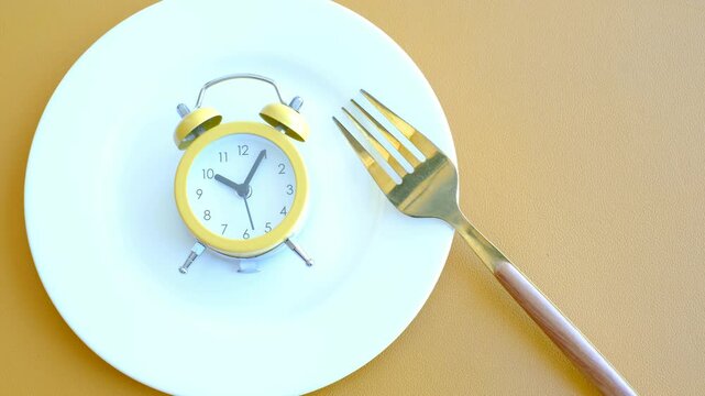 Empty plate with alarm clock and fork on brown background, intermittent fasting concept.
