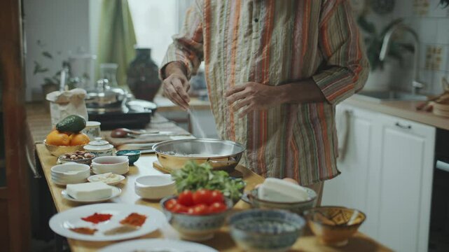 Senior Indian chef in turban and kurta adding salt to metal bowl and explaining recipe for dish as giving online cooking class or filming culinary vlog in cozy kitchen