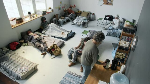 Wide high-angle surveillance camera shot of people displaced by natural disaster, flood or fire resting in emergency shelter &ndash; sleeping, pouring tea, listening to music, mom reading book to son