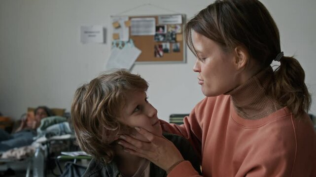 Medium shot of young Caucasian woman and 9-year-old son staying in emergency refugee accommodation, having escaped from fire, flood or war, comforting and kissing boy on forehead