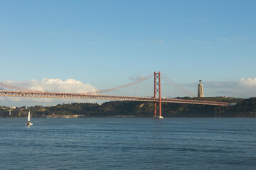 Naklejka premium A stunning view of the 25 de Abril Bridge in Lisbon, Portugal, spanning the Tagus River with the Cristo Rei monument in the background. A sailboat glides through the water under a clear blue sky.