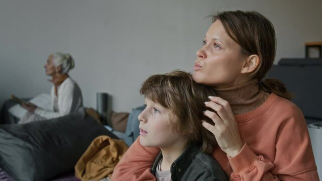 Medium closeup of loving young Caucasian refugee mother consoling son traumatized by war or natural disaster, talking and hugging, while resting in makeshift emergency shelter at daytime