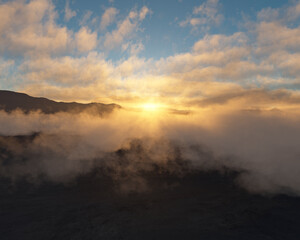 Clouds drift through a serene mountain landscape partially obscuring a rising or setting sun. The sky transitions from light blue to yellow and orange hues casting a warm glow across the scene.