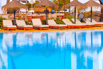 Poolside area with beige lounge chairs arranged in a row under thatched umbrellas, reflecting in blue water. Setting appears to be relaxing outdoor space of resort, designed for leisure and comfort
