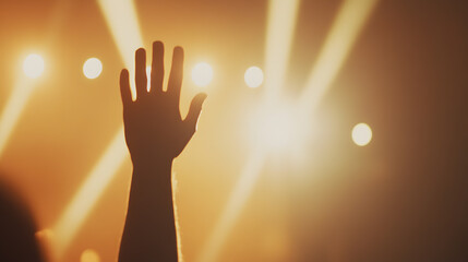 A close-up of hands waving in the air at a summer concert, bathed in warm, golden light. The stage glows in the background, slightly blurred, with beams of neon and LED lights cutting through the