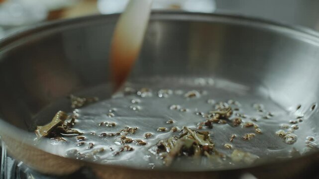 Close-up view of unrecognizable chef using spatula to stir whole Indian spices such as bay leaves and cumin seeds in hot oil inside frying pan, releasing aroma and flavor for cooking