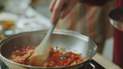 Young woman stirring chopped onion, tomatoes and spices in frying pan, learning Indian cuisine under guidance of senior chef in turban during culinary class in kitchen. Tilt-up, close-up shot