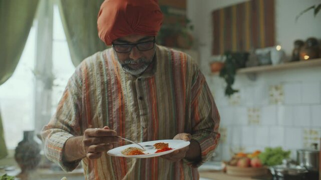 Senior Indian food blogger in turban and kurta showing aromatic spices on plate, describing recipe on camera and adding ingredients to pan on stove while cooking in kitchen