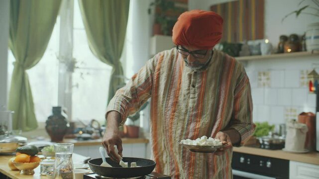 Indian food blogger in turban and kurta holding plate of cubed paneer cheese, speaking on camera and adding ingredients to preheated frying pan on stove as filming video recipe in kitchen