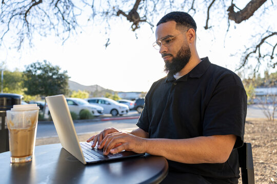 A confident man sits focused on his laptop, sipping on a delicious coffee in an inviting outdoor setting, surrounded by vibrant trees and passing cars in the background, creating a lively atmosphere - Powered by Adobe