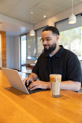 A man sits at a rustic wooden table in a cozy cafe, typing on his laptop while enjoying iced coffee. The large windows create a warm atmosphere for working, socializing, or relaxing