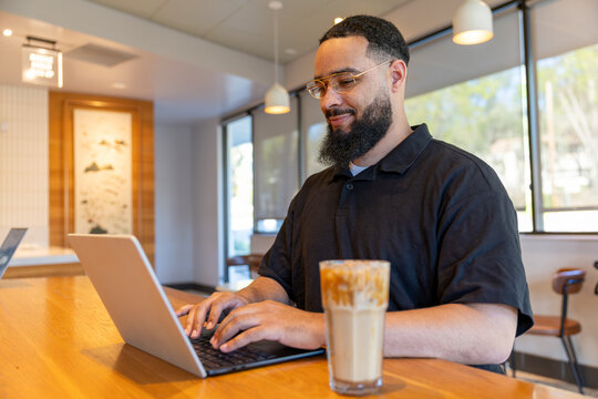 In a modern cafe, a man with a wellgroomed beard works on his laptop while enjoying iced coffee. The bright decor fosters a productive atmosphere, ideal for concentration and creativity