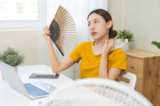 Suffering summer heat wave stroke, hot weather, tired asian young woman using handheld fan waving with blowing and holding cool water bottle on neck when working, ventilation when temperature high.