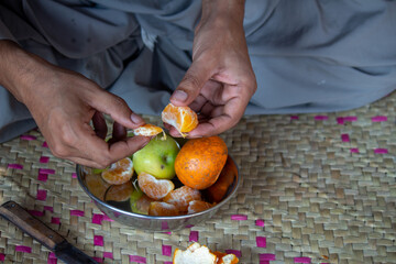 A person peeling a fresh orange with hands, stainless steel bowl with apples and oranges on a woven mat, depicting a rural lifestyle.


