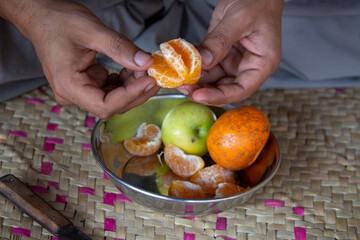 A person peeling a fresh orange with hands, stainless steel bowl with apples and oranges on a woven mat, depicting a rural lifestyle.

