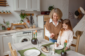 Elderly woman grandmother and little granddaughter joyfully set the table at home in the kitchen, learn etiquette