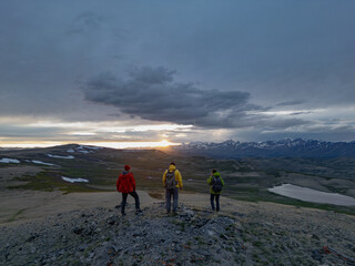 Obraz premium Friends of tourists and travelers stand on the top of the mountain during sunset and look into the distance at the snowy mountains. People in bright clothes in the mountains during sunset