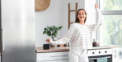 Beautiful young happy woman with wireless portable speaker in kitchen at home